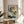 May include: Japanese woodblock print in wooden frame showing white cranes by pine tree with orange sun and blue-green foliage, displayed on round wooden side table next to beige upholstered chair, with plant leaves visible in corner and natural sunlight casting shadows on white wall