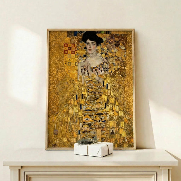 May include: Ornate golden portrait painting in light wood frame displayed on white wooden console table with small wrapped gift box. The artwork features a woman in elaborate patterned dress surrounded by intricate geometric designs and swirling motifs in rich golds, yellows and earth tones. The decorative Art Nouveau style shows detailed ornamental elements throughout the composition against a luxurious golden background.