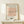 May include: Framed art print displaying minimalist line drawing of a dachshund dog in continuous black lines on peachy-pink background, with "PICASSO" and "LE CHIEN" text above and below. Natural wood frame sits on light wooden shelf against cream wall, with small white wrapped gift beside it.