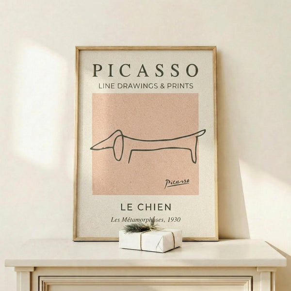 May include: Framed art print displaying minimalist line drawing of a dachshund dog in continuous black lines on peachy-pink background, with "PICASSO" and "LE CHIEN" text above and below. Natural wood frame sits on light wooden shelf against cream wall, with small white wrapped gift beside it.