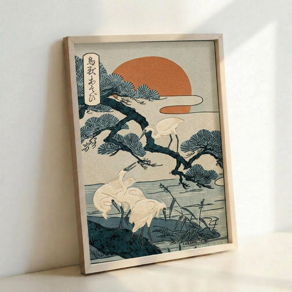 May include: Framed Japanese woodblock print showing white cranes near water with a gnarled pine tree, orange sun, and Japanese text in upper left corner. The artwork features traditional ukiyo-e style with blue-green tones and cream background, displayed in light wood frame leaning against white wall.