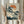 May include: Person in cream ruffled sleeves holding framed Japanese woodblock print featuring white cranes by water with pine tree, orange sun, and stylized clouds. Print has beige mat in light wood frame. Dried pampas grass visible beside figure against soft curtained background.