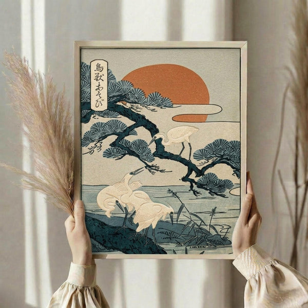 May include: Person in cream ruffled sleeves holding framed Japanese woodblock print featuring white cranes by water with pine tree, orange sun, and stylized clouds. Print has beige mat in light wood frame. Dried pampas grass visible beside figure against soft curtained background.