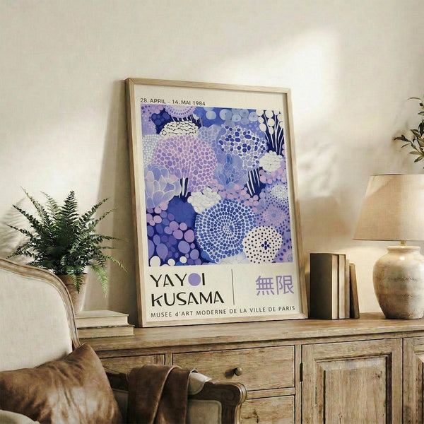 A framed poster of Yayoi Kusama's artwork hangs on a wall in a living room, surrounded by various decorative items such as a potted plant, books, and a lamp.