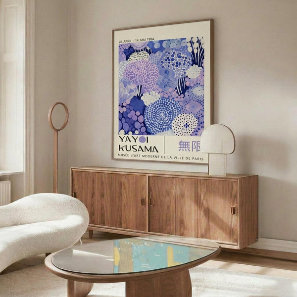 a modern living room with a large abstract art print hanging on the wall above a wooden cabinet. The print has a vibrant blue and purple color scheme with various shapes and patterns. The room also includes a white sofa, a glass coffee table, and a lamp on a side table.