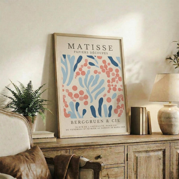 a framed poster of the M.C. Escher's "Paperiers D'Ecoques" artwork, which is a collection of various papers and papers arranged in a collage-like manner. The poster is displayed on a wooden cabinet or shelf, with a lamp and a plant nearby.