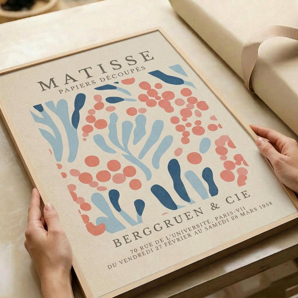 a framed poster of the Matisse paper art print titled "Papiers decoupes" (Paper Decoupes) with a floral pattern in shades of blue, red, and orange. The poster is being held by a person's hands, and there is a beige gift box visible in the background.