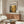 a modern living room with a large wooden cabinet, a white sofa, and a glass coffee table. A framed poster with a gold and black abstract design is hanging on the wall above the cabinet.