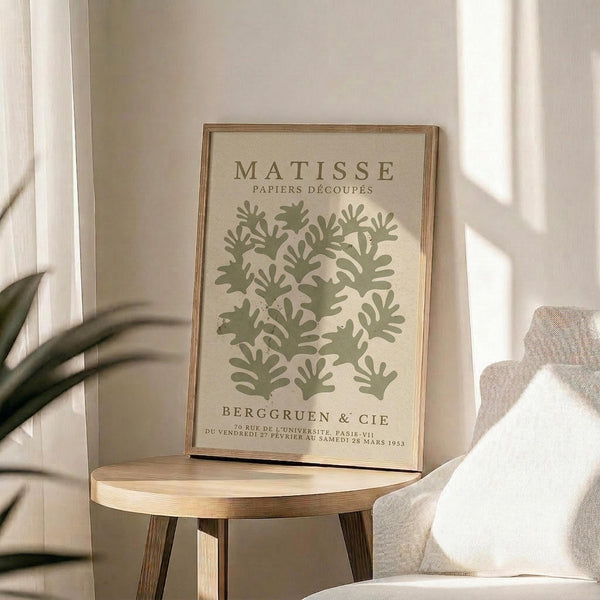 a framed poster of the famous painting "Matsisse" by the artist Henri Matisse, displayed on a wooden table in a room with a white chair and a plant in the background.