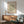 a living room with a framed poster of a leafy green abstract artwork hanging on the wall above a wooden cabinet. The cabinet has a white lamp on top of it. There is a green armchair in the foreground, and the room has a wooden floor.