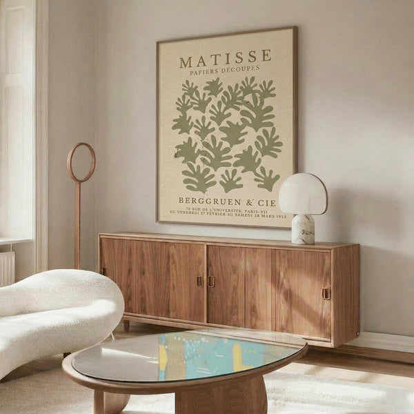 a living room with a large framed poster of a green leafy pattern on the wall, a wooden cabinet, a white sofa, and a glass-topped coffee table.