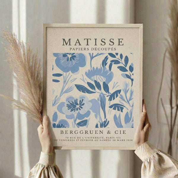 A person is holding up a framed poster of the Métisse papiers decoupes artwork by Berg Gruen & Cie, which features a floral design in blue and white colors.