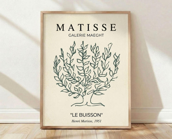 May include: Framed botanical line drawing print in light wood frame, featuring black outlined bush or shrub design on cream background. Text reads "MATISSE GALERIE MAEGHT" at top and "LE BUISSON Henri Matisse, 1951" at bottom. Frame sits on wooden floor against white wall with soft natural lighting creating gentle shadows.