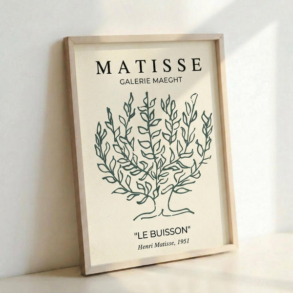 May include: Framed art print featuring a minimalist line drawing of a leafy bush or shrub in dark green on cream background. Text reads "MATISSE GALERIE MAEGHT" at top and "LE BUISSON" with "Henri Matisse, 1951" at bottom. Simple botanical illustration in characteristic Matisse style, displayed in light wooden frame leaning against white wall with natural lighting creating soft shadows.
