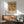 May include: Framed vintage botanical poster with ornate brown and cream floral pattern featuring large blooms and flowing stems, displayed above mid-century modern wood credenza with white ceramic lamp. Green upholstered armchair with wooden frame sits nearby in bright, minimalist living room with white walls and natural sunlight streaming across the scene.