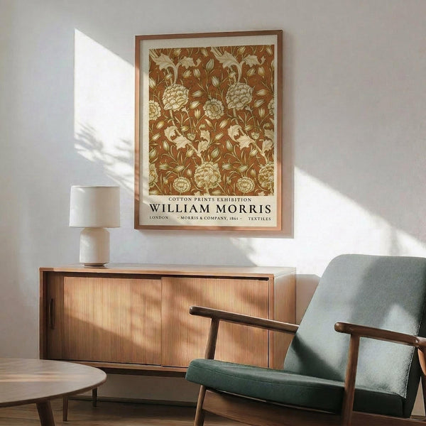 May include: Framed vintage botanical poster with ornate brown and cream floral pattern featuring large blooms and flowing stems, displayed above mid-century modern wood credenza with white ceramic lamp. Green upholstered armchair with wooden frame sits nearby in bright, minimalist living room with white walls and natural sunlight streaming across the scene.