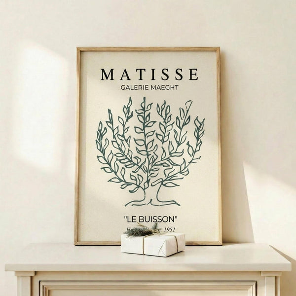May include: Framed art print displaying a simple line drawing of a leafy bush or tree in dark green on cream background, with "MATISSE GALERIE MAEGHT" and "LE BUISSON" text above and below. Light wood frame sits on white wooden shelf against cream wall, with small wrapped gift box nearby.