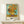 May include: Van Gogh's famous sunflowers painting reproduction in thin wooden frame, displayed on light wood shelf against white wall. Vibrant yellow sunflowers in ceramic vase against turquoise background, with "VAN GOGH" and "1888" text below artwork. Small white wrapped gift box with green sprig sits on shelf next to frame.