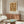 May include: Framed botanical print with rust-colored floral pattern hanging above mid-century walnut credenza in minimalist living room. White curved chair and oval glass coffee table with blue and yellow art book visible. Warm wood tones complement the vintage-style poster's earthy palette.