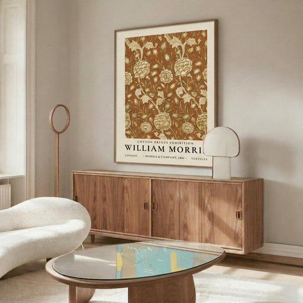 May include: Framed botanical print with rust-colored floral pattern hanging above mid-century walnut credenza in minimalist living room. White curved chair and oval glass coffee table with blue and yellow art book visible. Warm wood tones complement the vintage-style poster's earthy palette.