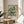 May include: Framed Van Gogh floral art print displayed on round wooden side table next to beige upholstered chair. Print shows white roses in vase with green brushstroke background in light wood frame. Soft natural lighting creates shadows on cream wall, with plant leaves visible in corner of bright, minimalist room setting.