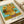 May include: Van Gogh's Sunflowers reproduction print displayed on white paper with cream ribbon nearby. The artwork shows vibrant yellow sunflowers in a ceramic vase against turquoise and olive green background. Print features white border with "VAN GOGH" and "Sunflowers, 1888" text at bottom. Visible brushstroke texture and warm golden tones throughout the classic Post-Impressionist painting reproduction.