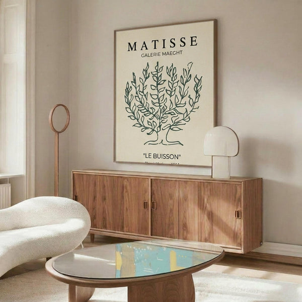 May include: Matisse botanical art print in thin wooden frame hanging above mid-century walnut credenza in modern living room. Cream-colored poster features hand-drawn green bush illustration with gallery text. Room has white curved chair, glass coffee table, white mushroom lamp, and brass floor lamp against beige walls.