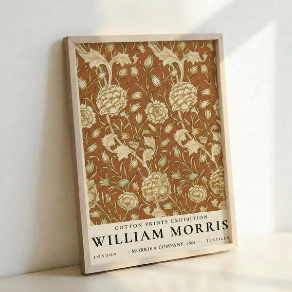 May include: Framed poster with William Morris floral textile pattern in warm brown and cream tones, featuring ornate blooming flowers, curved stems and decorative leaves. Wooden frame leans against white wall, showing exhibition-style text at bottom. Arts and Crafts movement design with intricate botanical motifs in vintage color palette.
