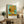 May include: Framed Van Gogh Sunflowers reproduction print displayed on rustic wooden sideboard in neutral-toned living room. Yellow sunflowers in vase against turquoise background with artist name below. Scene shows beige armchair with brown throw pillow, potted fern, stacked books, ceramic vases, and white walls with natural lighting.
