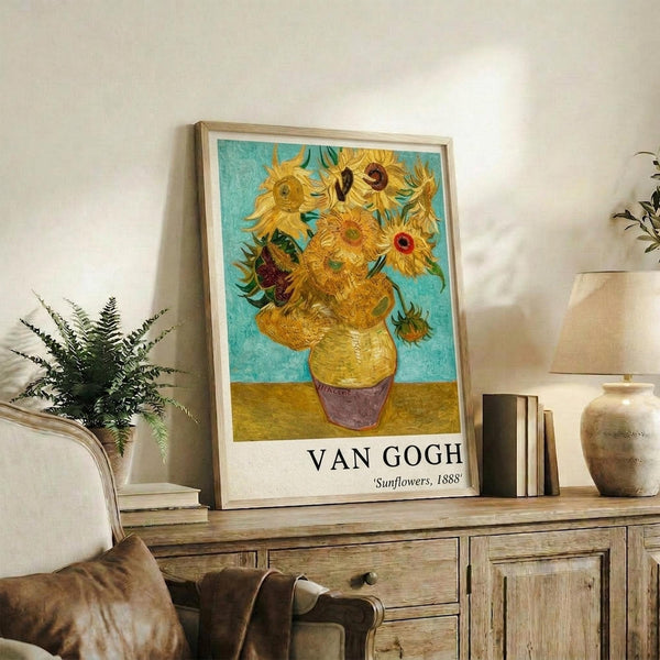 May include: Framed Van Gogh Sunflowers reproduction print displayed on rustic wooden sideboard in neutral-toned living room. Yellow sunflowers in vase against turquoise background with artist name below. Scene shows beige armchair with brown throw pillow, potted fern, stacked books, ceramic vases, and white walls with natural lighting.