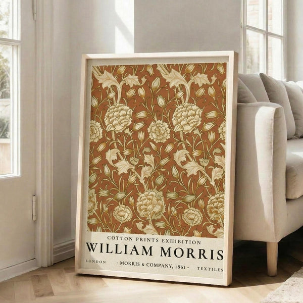 May include: Framed poster with William Morris floral pattern in warm brown and cream tones, featuring detailed botanical motifs with large blooms and flowing leaves. Exhibition-style text at bottom reads "Cotton Prints Exhibition William Morris London Morris & Company 1861 Textiles". Light wooden frame leans against white wall next to beige upholstered chair in bright room with tall windows.