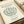 May include: Cream-colored art print with minimalist line drawing of a leafy bush or plant in sage green. Features "MATISSE" and "GALERIE MAEGHT" at top, "LE BUISSON" and "Henri Matisse, 1951" at bottom. Print has thin border frame, shown on neutral background with beige ribbon nearby.