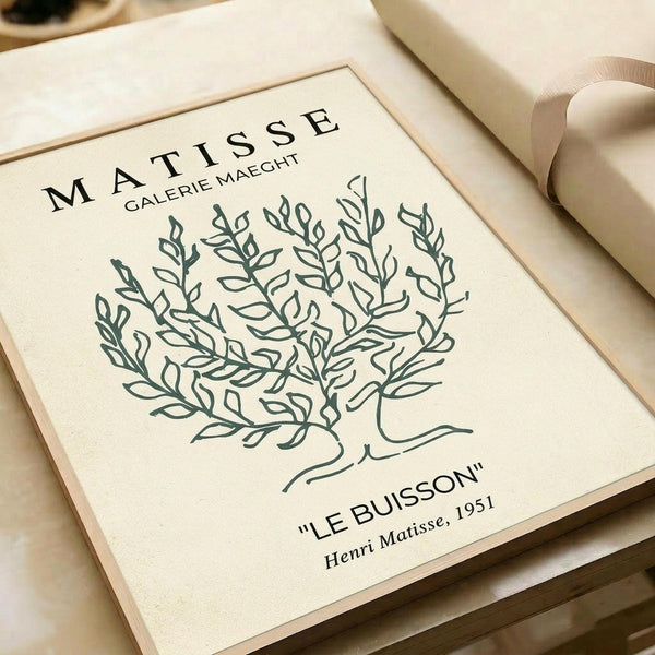 May include: Cream-colored art print with minimalist line drawing of a leafy bush or plant in sage green. Features "MATISSE" and "GALERIE MAEGHT" at top, "LE BUISSON" and "Henri Matisse, 1951" at bottom. Print has thin border frame, shown on neutral background with beige ribbon nearby.