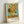 May include: Framed reproduction of Van Gogh's famous sunflowers painting in natural wood frame, leaning against white wall. Artwork shows vibrant yellow sunflowers in ceramic vase against turquoise background, with text reading "VAN GOGH 'Sunflowers, 1888'" at bottom. Visible brushstroke texture and warm golden tones throughout the floral still life composition.
