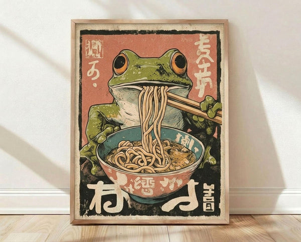 May include: Framed vintage-style Japanese poster showing a green frog with orange eyes eating ramen noodles with chopsticks from a red and teal bowl, set against a coral pink background with Japanese text, displayed in a light wood frame leaning against a white wall on light wooden flooring.