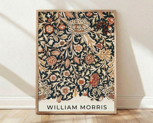 May include: Framed botanical print with intricate dark navy background featuring ornate floral patterns in cream, coral, and gold tones. The design shows detailed flowers, leaves, and decorative motifs in William Morris's signature Arts and Crafts style. Light wood frame sits on wooden floor against white wall with soft natural lighting creating gentle shadows.