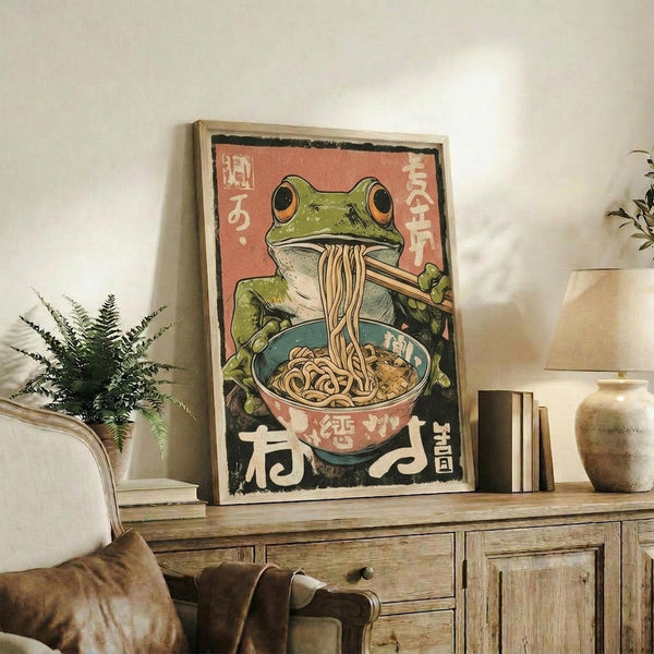 May include: Framed vintage-style Japanese poster featuring a green frog with large orange eyes eating noodles from a red and teal ramen bowl, holding chopsticks, with Japanese text on coral pink background. Wooden frame displayed on rustic wood sideboard next to fern plant, books, and ceramic vase in modern living room setting with beige chair visible.