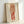May include: Framed art print leaning against white wall showing expressionist-style drawing of standing figure in reddish-orange tones. Figure wears loose draped clothing on upper body with bare red-tinted legs below. Artwork has cream background with artist signature in corner, displayed in simple light wood frame on white surface.
