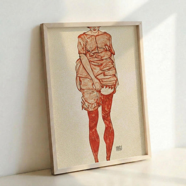 May include: Framed art print leaning against white wall showing expressionist-style drawing of standing figure in reddish-orange tones. Figure wears loose draped clothing on upper body with bare red-tinted legs below. Artwork has cream background with artist signature in corner, displayed in simple light wood frame on white surface.