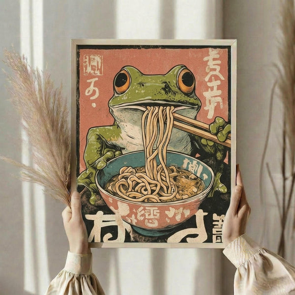 May include: Person holding framed vintage-style Japanese poster featuring green frog with orange eyes eating noodles from a ramen bowl using chopsticks, with Japanese text on coral-colored background. Light wooden frame, held against white curtained window with dried wheat stems visible to the left.