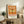 May include: Orange vintage-style museum poster featuring a detailed dragonfly illustration with Japanese and English text, displayed in a light wood frame on a rustic wooden sideboard alongside a fern plant, stacked books, and ceramic pottery in a warm, neutral-toned living room setting.