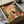 May include: Framed illustration of a black cat sitting on a toilet reading a newspaper in a coral pink tiled bathroom with yellow lighting, tropical plants, and mid-century modern styling. The artwork is packed in a cardboard shipping box with protective brown paper wrapping, surrounded by white tissue paper on a textured surface.