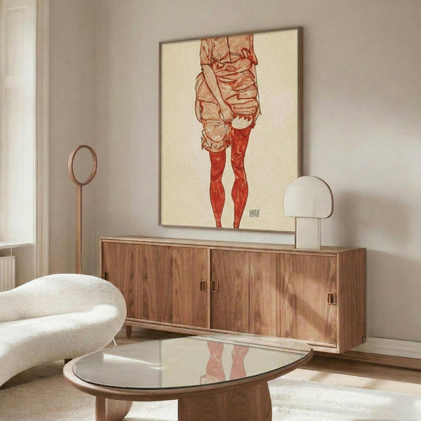 May include: Framed art print featuring a figure study in red and beige tones hanging above a wood credenza in a modern living room. The artwork shows partial human form rendered in warm reddish hues against cream background. Room styled with white curved chair, glass coffee table, and contemporary lighting fixtures on neutral walls.
