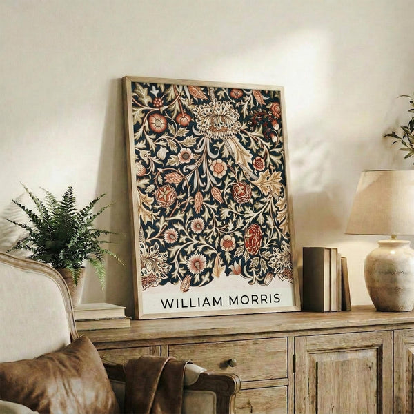 May include: Framed botanical art print with intricate dark navy background featuring ornate floral patterns in cream, coral, and gold tones. The vintage-style design shows detailed leaves, roses, and decorative motifs. Display shows rustic wooden sideboard with books, ceramic vases, fern plant, and beige upholstered chair in warm, neutral-toned room setting.