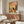 May include: Colorful illustration of black cat sitting on toilet reading newspaper in vibrant pink and yellow tiled bathroom with plants and shelving, displayed as framed artwork above mid-century walnut credenza in modern living room with white sculptural chair, round glass coffee table, and contemporary lighting fixtures