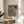 May include: Framed botanical poster with intricate dark navy background featuring ornate floral pattern in coral, cream and gold tones, displayed on round wooden side table next to beige upholstered chair, with plant leaves visible and natural sunlight streaming across white walls
