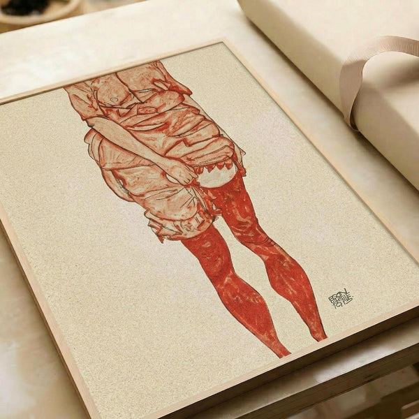 May include: Framed art print showing an expressionist figure drawing in red and beige tones, depicting a person in flowing garments with elongated limbs in an emotional pose. The artwork appears mounted in a light wooden frame, positioned on a cream-colored surface next to rolled paper tubes, creating an artistic workspace setting with warm, muted lighting.