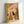May include: Framed illustration print leaning against white wall showing a black cat sitting on a toilet reading a newspaper in a colorful bathroom with pink and coral tiled walls, yellow shower curtain, potted plants, small framed artwork, and checkered floor tiles in a whimsical, stylized art style with warm lighting.