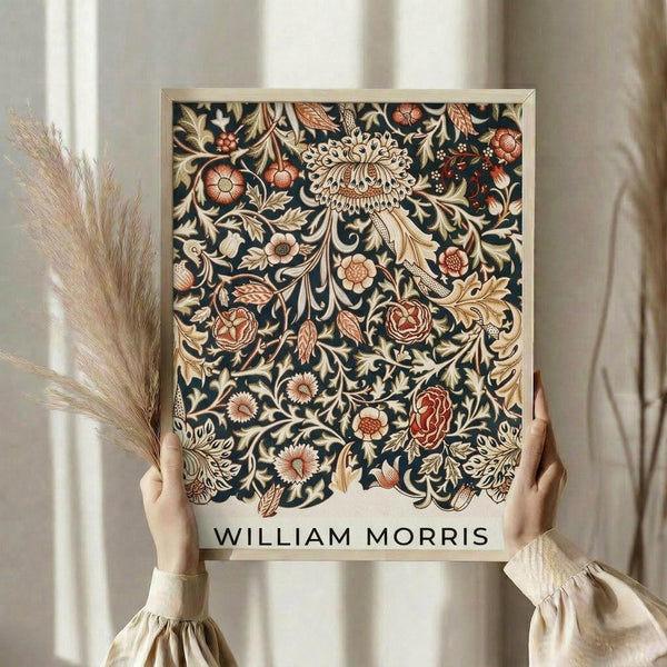May include: Person in cream clothing holding framed botanical art print featuring intricate dark blue background with ornate floral pattern in rust, coral, and cream tones. Decorative wheat stalks visible alongside neutral curtained interior setting with natural lighting.