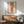 May include: Framed artwork showing a figure in loose clothing with red stockings, displayed above a wooden sideboard with white lamp in modern living room setting with sage green armchair and natural lighting creating wall shadows